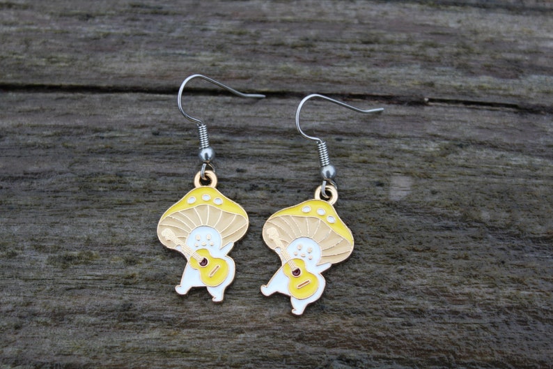 May include: A pair of whimsical mushroom-shaped earrings. Each earring features a yellow and tan mushroom cap with a smiling face and a small guitar. The earrings have silver-colored hooks and are set against a wooden background.
