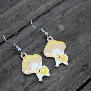 May include: A pair of whimsical mushroom-shaped earrings. Each earring features a yellow and tan mushroom cap with white spots, and a white body playing a yellow ukulele. The earrings have silver-tone hooks and a gold-tone accent.