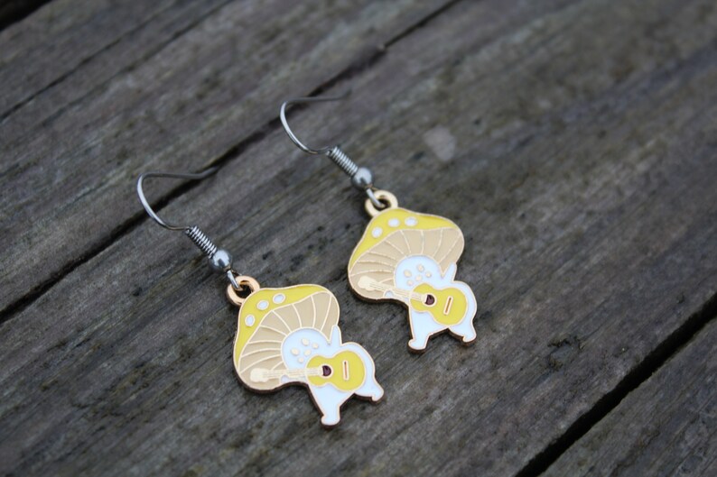 May include: A pair of whimsical mushroom-shaped earrings. Each earring features a yellow and tan mushroom cap with white spots, and a white body playing a yellow ukulele. The earrings have silver-tone hooks and a gold-tone accent.