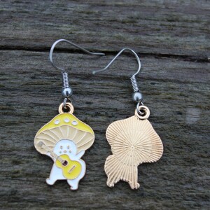 May include: A pair of mushroom-shaped earrings. Each earring features a yellow and beige mushroom design with a white figure playing a yellow guitar. The earrings have silver-colored hooks.