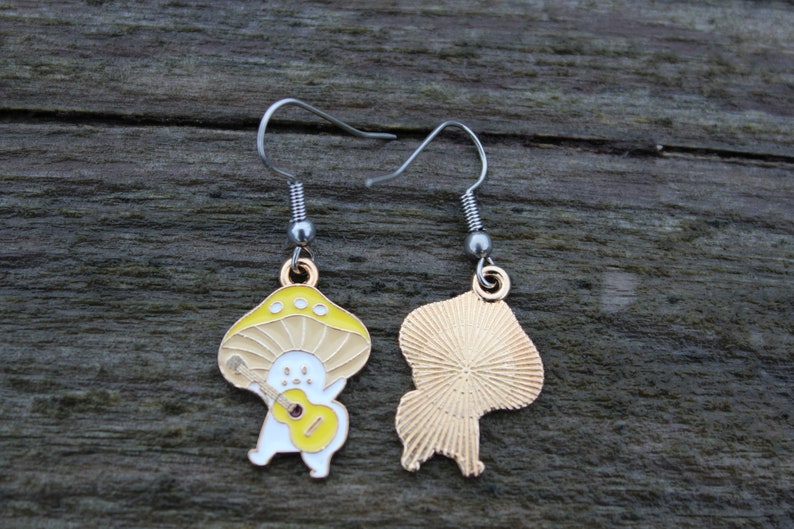 May include: A pair of mushroom-shaped earrings. Each earring features a yellow and beige mushroom design with a white figure playing a yellow guitar. The earrings have silver-colored hooks.
