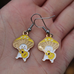 May include: A pair of whimsical mushroom earrings. Each earring features a yellow and tan mushroom cap with a white body playing a yellow guitar. The earrings have silver-colored hooks.