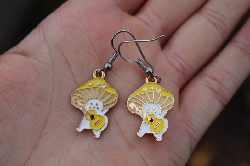May include: A pair of whimsical mushroom earrings. Each earring features a yellow and tan mushroom cap with a white body playing a yellow guitar. The earrings have silver-colored hooks.