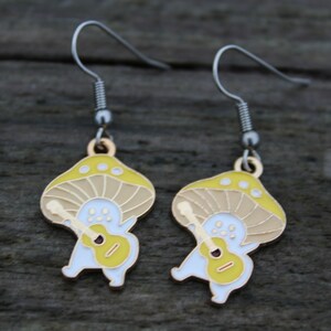 May include: A pair of whimsical mushroom-shaped earrings. Each earring features a yellow and tan mushroom cap with a white body playing a yellow guitar. The earrings have silver-colored hooks.