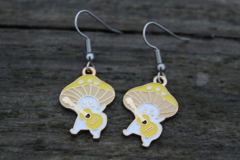 May include: A pair of whimsical mushroom-shaped earrings. Each earring features a yellow and tan mushroom cap with a white body playing a yellow guitar. The earrings have silver-colored hooks.