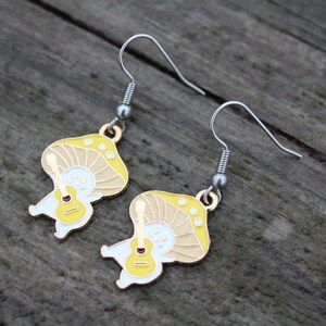 May include: A pair of whimsical mushroom earrings. Each earring features a yellow and tan mushroom design with a white body holding a small guitar. The earrings have silver-colored hooks and are set against a wooden background.