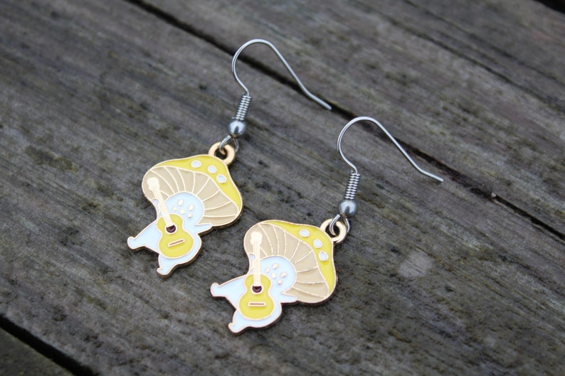 May include: A pair of whimsical mushroom earrings. Each earring features a yellow and tan mushroom design with a white body holding a small guitar. The earrings have silver-colored hooks and are set against a wooden background.