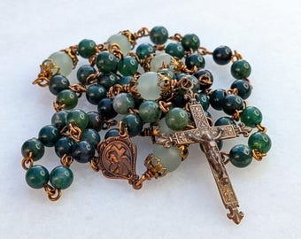 Könnte beinhalten: Ein Rosenkranz mit grünen und klaren Perlen und einem Bronzekreuz. Die Perlen sind eine Mischung aus dunklem und hellem Grün, mit einigen klaren Perlen. Das Kruzifix und die Verbindungsteile sind aus Bronze.