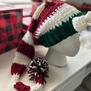 May include: A handmade crochet elf hat in red, white, and green. The hat features a long, curved tip and a pom-pom at the end. The hat is displayed on a white mannequin head. The hat is a festive accessory.