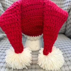 May include: A handmade, red crochet hat with long, pointed ends and white pom-poms. The hat has a white, scalloped trim at the base. This festive hat is perfect for costumes or holiday celebrations.