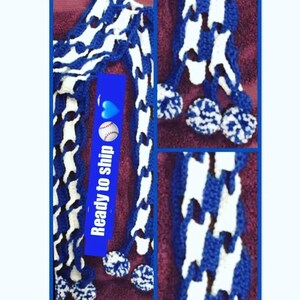 May include: A blue and white crocheted scarf with a unique pattern and pom-poms at the ends. The scarf is displayed on a maroon surface. The image also includes a blue rectangle with the words "Ready to ship" and a baseball graphic.