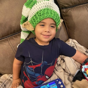 May include: A child wearing a handmade crochet elf hat with green and white stripes and tan ears. The child is wearing a navy blue Spider-Man t-shirt and holding a blue handheld gaming device.