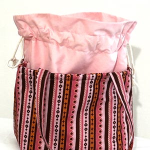 May include: A pink and patterned tote bag with a drawstring closure. The outer bag features vertical stripes in shades of pink, brown, and orange. The inner bag is a solid light pink. The bag has a long strap for carrying.