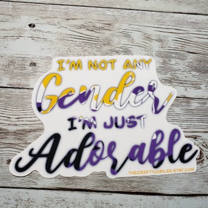 May include: A white sticker with the text "I'm not any gender, I'm just adorable" in black and purple lettering. The text is surrounded by a yellow, white, and purple design.