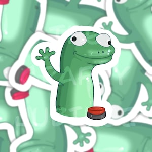 May include: A green cartoon gecko sticker with a friendly expression, waving a hand. The sticker features a red and black button at the bottom. The background is a light green, with other gecko stickers visible.