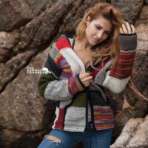 May include: A woman wearing a multi-coloured patchwork cardigan with a hood and long sleeves. The cardigan is made of different types of knit fabric and has a zipper closure. The woman is wearing blue jeans and has long, wavy hair.
