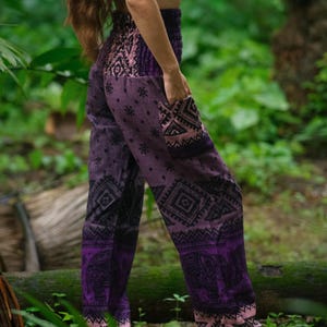 May include: High-waisted purple pants featuring a black elephant print, geometric patterns, and a small pocket on the side. The pants have a smocked elastic waistband and a loose, relaxed fit with a tapered leg design in shades of violet and black.