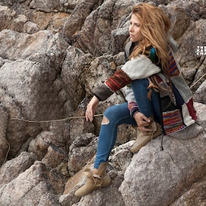 May include: A woman wearing a colorful patchwork cardigan, blue jeans, and brown boots sits on a rocky outcrop. The text "THE KARMIC CHAMELEON" is visible in the upper right corner of the image.