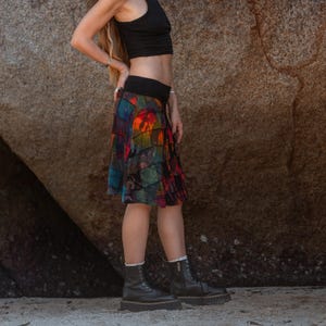 Puede incluir: Una falda colorida, hasta la rodilla, con un diseño escalonado. La falda presenta un vibrante estampado tie-dye en tonos de rojo, naranja, azul y verde. La cintura es negra. La persona lleva botas negras.