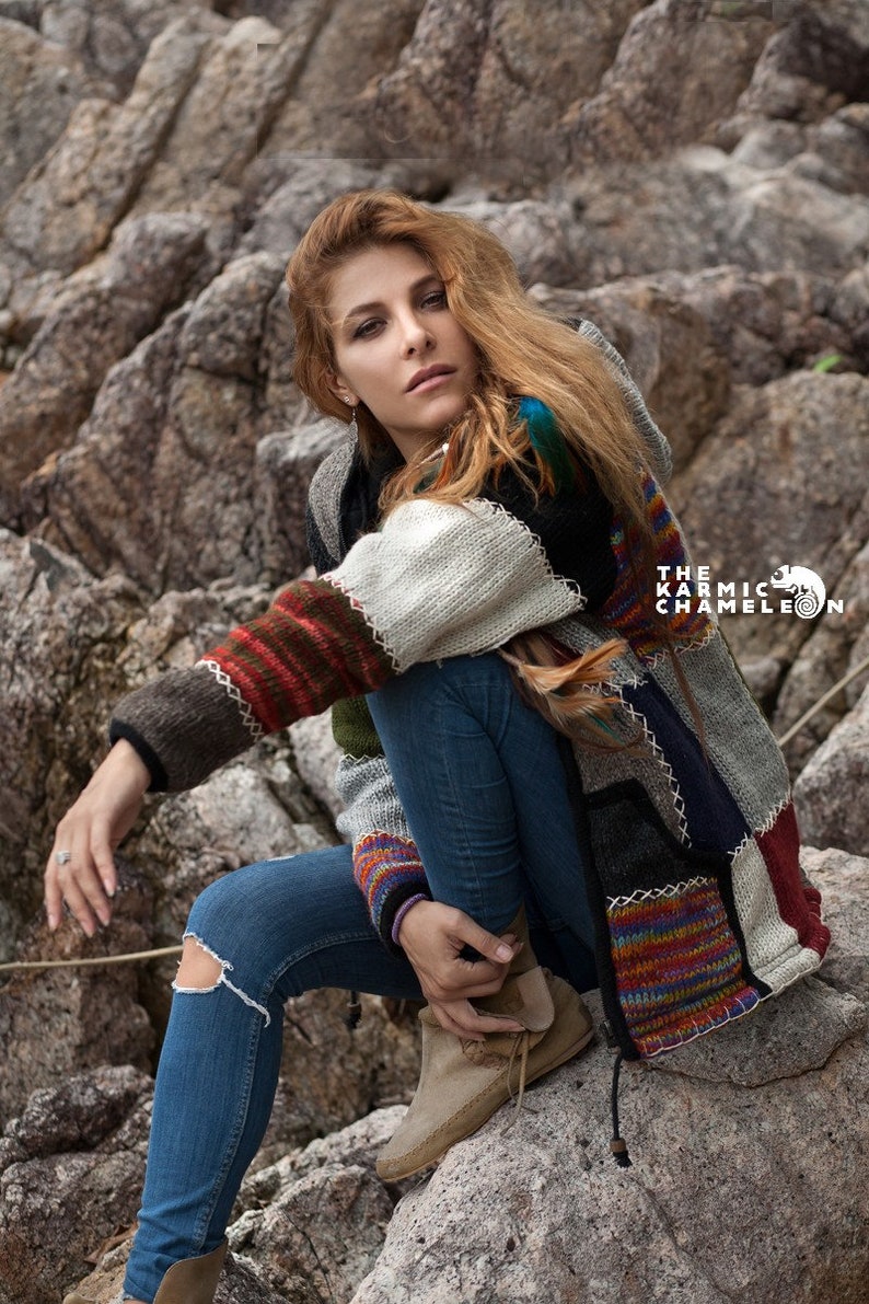 May include: A woman wearing a multi-colored patchwork cardigan with a hood, blue jeans, and brown suede boots. The cardigan has a variety of colors and textures, including brown, white, red, blue, and green. The woman is sitting on a rock with a rocky background.