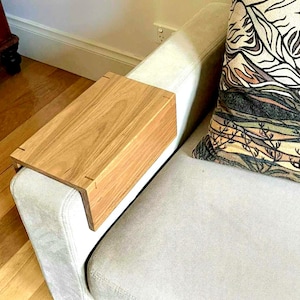 Könnte beinhalten: Eine rechteckige Armlehnenablage aus Holz mit hellbrauner Holzmaserung, die über die Armlehne eines Sofas gelegt wird. Die Ablage weist sichtbare Verbindungsdetails an den Ecken sowie eine glatte, flache Oberfläche zum Abstellen auf.