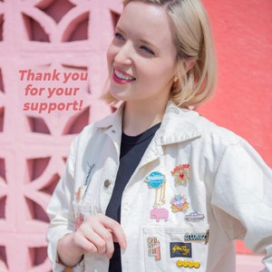 Puede incluir: Una mujer con cabello rubio sonr&iacute;e frente a una pared rosa. Lleva una chaqueta blanca con una variedad de pines, incluyendo un elefante rosa, un globo azul y dorado, y un pin rojo y blanco "Lucky".