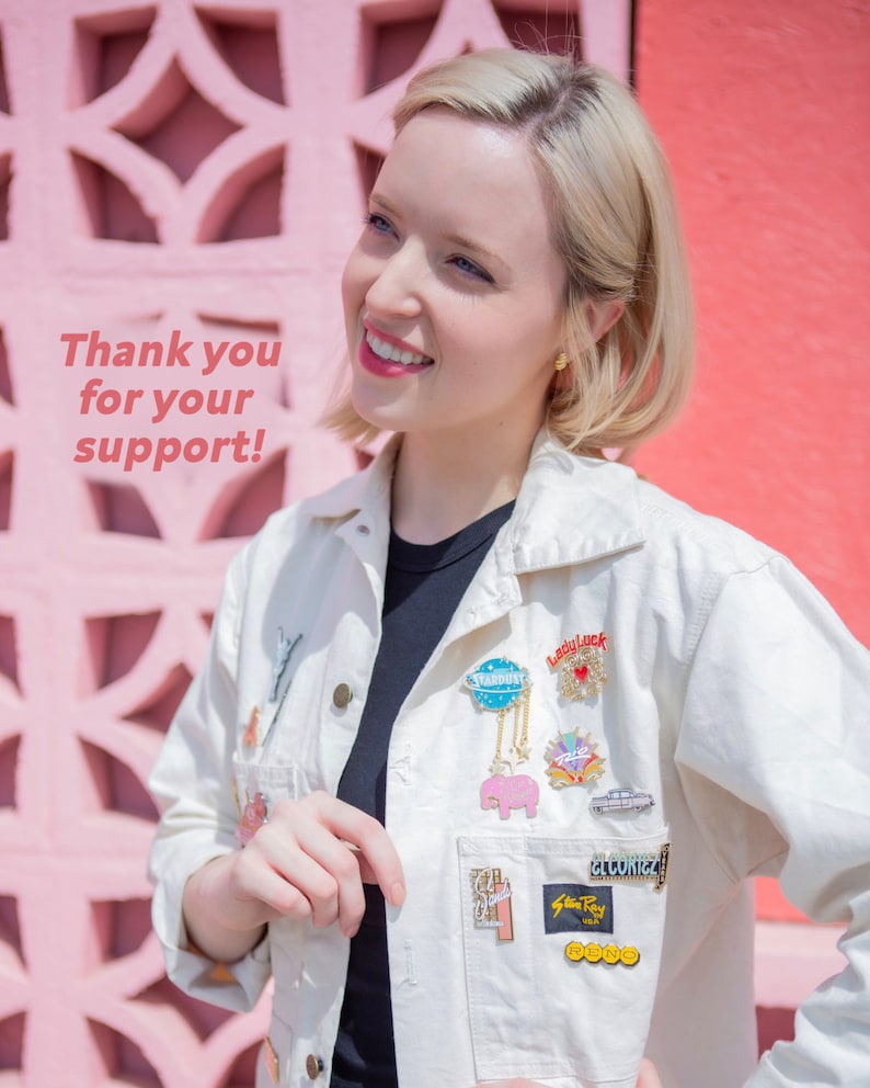 Puede incluir: Una mujer con cabello rubio sonr&iacute;e frente a una pared rosa. Lleva una chaqueta blanca con una variedad de pines, incluyendo un elefante rosa, un globo azul y dorado, y un pin rojo y blanco "Lucky".