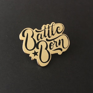 May include: Gold-colored metal pin with a black outline of the words "Battle Born" and a small star in the center.