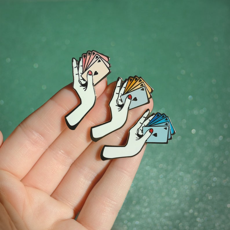 Puede incluir: Tres pines de esmalte que representan una mano sosteniendo un abanico de cartas de juego. Las cartas son de diferentes colores: rosa, amarillo y azul. Cada pin tiene un coraz&oacute;n negro en la carta.