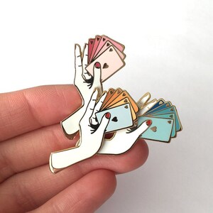 Puede incluir: Pin de esmalte dorado con dos manos sosteniendo un abanico de cartas de juego. Las cartas son en tonos de rosa, naranja y azul.