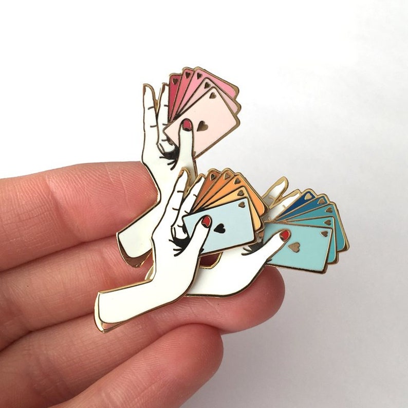 Puede incluir: Pin de esmalte dorado con dos manos sosteniendo un abanico de cartas de juego. Las cartas son en tonos de rosa, naranja y azul.
