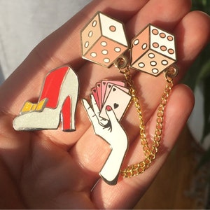 Puede incluir: Un conjunto de tres pines de esmalte que representan un zapato blanco de tac&oacute;n alto, una mano que sostiene cartas de juego y dos dados rosas conectados por una cadena dorada.