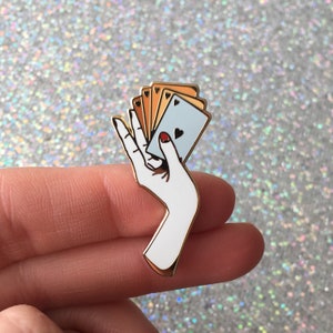 Puede incluir: Pin de esmalte dorado con una mano blanca sosteniendo un abanico de cartas de juego. La carta superior es un as de corazones azul.