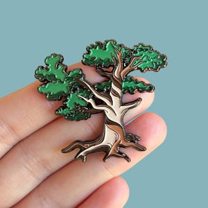May include: A metal enamel pin shaped like a bonsai tree with a light tan trunk and dark brown outlines. The tree features vibrant green foliage sections with dark brown borders, creating a stylized representation of a small, twisted tree.