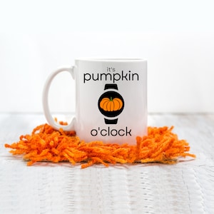 Puede incluir: Taza de cerámica blanca con un gráfico negro de una calabaza dentro de una esfera de reloj y el texto "it's pumpkin o'clock". La taza está sobre un posavasos de pelo naranja brillante.