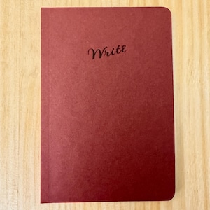 May include: A dark red notebook with a textured cover featuring the word Write printed in black script font on the upper center. The notebook has a rectangular shape with rounded corners on the right side and a vertical spine crease on the left.