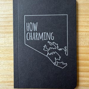 May include: A black notebook with a rectangular cover featuring the text HOW CHARMING printed in white. Below the text is a white line-art outline of a geographic map shape, positioned on the right side of the cover with a vertical spine.