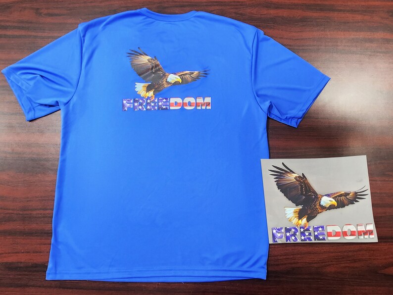May include: A blue t-shirt with a graphic of a bald eagle flying with the word "FREEDOM" printed in red, white, and blue.