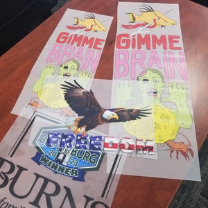 May include: Three iron-on transfers on a clear background. The top two transfers feature a zombie with the text "Gimme Brain" in pink and yellow. The bottom transfer features a bald eagle with the text "FREEDOM" in red, white, and blue.