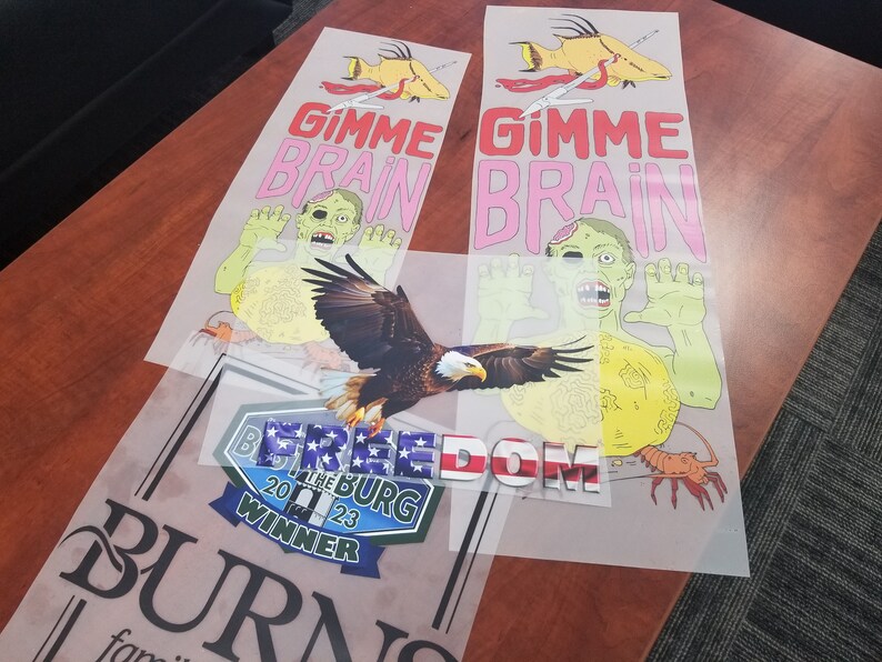 May include: Three iron-on transfers on a clear background. The top two transfers feature a zombie with the text "Gimme Brain" in pink and yellow. The bottom transfer features a bald eagle with the text "FREEDOM" in red, white, and blue.