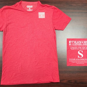 May include: Red short-sleeved t-shirt with a crew neck. The t-shirt has a tag that says "Black & Denim Apparel Company" and "Apparel for the Brave" in white text. The tag also says "Made in U.S.A. with 100% Cotton" and has a size "S" printed on it.