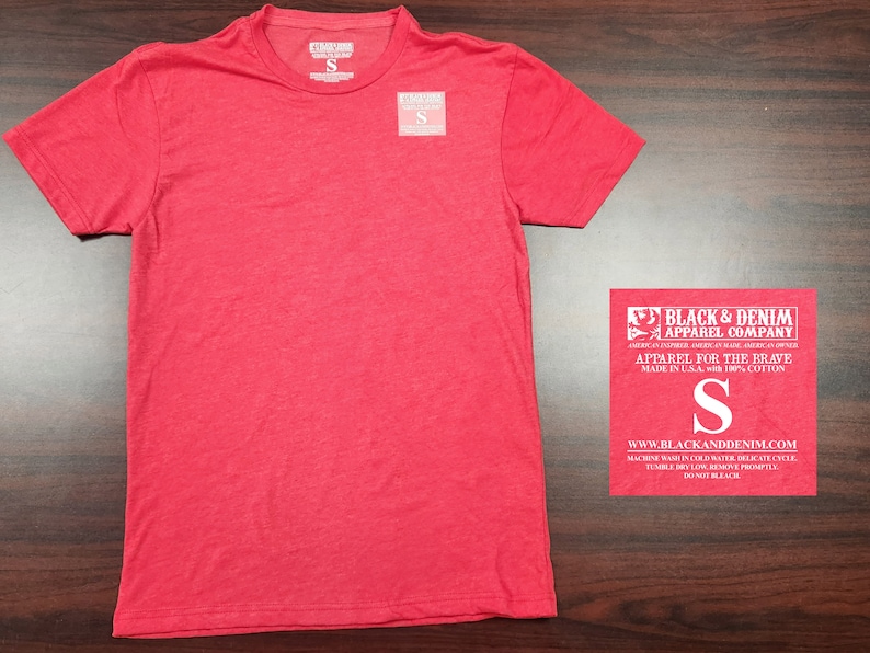 May include: Red short-sleeved t-shirt with a crew neck. The t-shirt has a tag that says "Black & Denim Apparel Company" and "Apparel for the Brave" in white text. The tag also says "Made in U.S.A. with 100% Cotton" and has a size "S" printed on it.