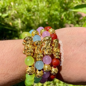 May include: A stack of five bracelets with gold charms and colorful beads. The beads are green, light blue, purple, and red. The charms are in the shape of a lion. The bracelets are stacked on a person's wrist. The text "@thegiftsofisis" is visible on the image.