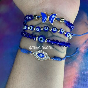 May include: Set of four blue evil eye bracelets with gold accents. One bracelet features a blue butterfly charm. The bracelets are on a person's wrist. @TheGiftsOfIsis
