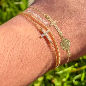 May include: A gold bracelet with a charm of the Virgin Mary and two cross charms with small diamonds. The bracelet is on a person's wrist.