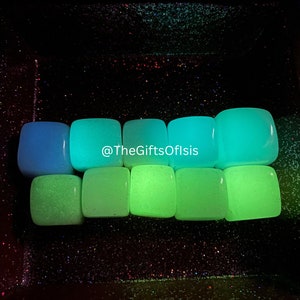 以下が含まれることがあります： 青と緑の色合いの、暗闇で光る正方形の石が8つ。石は、キラキラ光る黒い表面に一列に並んでいます。画像の中央には「@TheGiftsOfIsis」というテキストが表示されています。