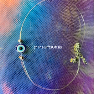 May include: A silver chain necklace with a blue evil eye charm. The chain is on a blue and purple background with a white grid pattern. The text "@TheGiftsOfIsis" is visible in the image.