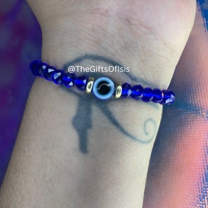 May include: A blue beaded bracelet with a gold evil eye charm. The bracelet is on a person's wrist. The text "@TheGiftsOfIsis" is visible on the bracelet.
