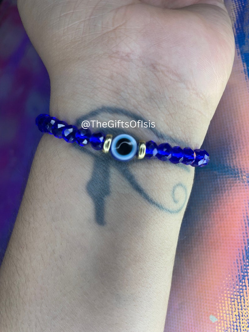 May include: A blue beaded bracelet with a gold evil eye charm. The bracelet is on a person's wrist. The text "@TheGiftsOfIsis" is visible on the bracelet.