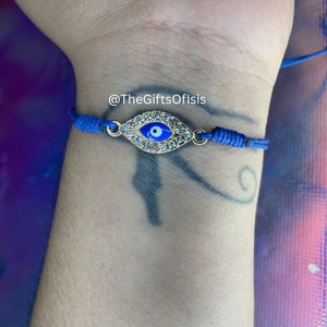 May include: Blue string bracelet with a gold evil eye charm with rhinestones. The charm has a blue eye with a white pupil. The bracelet is on a person's wrist. @TheGiftsOfIsis